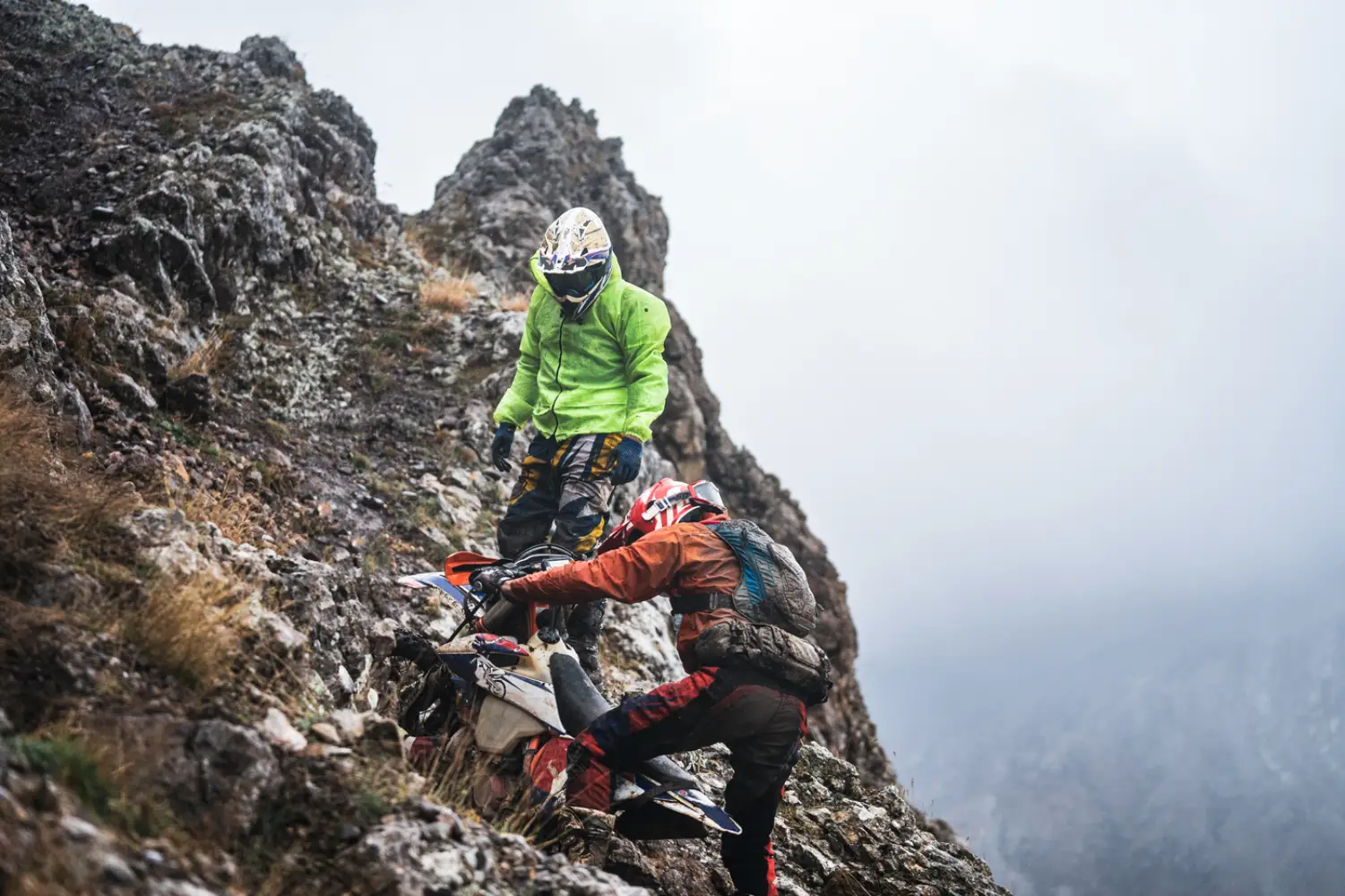 Two riders hauling a bike up a rocky ridge in the mist