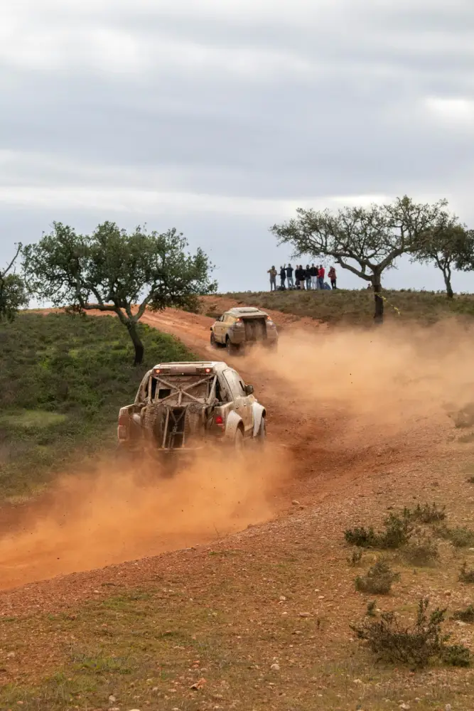 Rally-raid trucks racing up a dusty hill past spectators